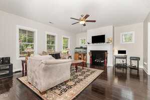 A living room with dark wood floors, beige walls, a patterned rug, two beige sofas, a fireplace with a TV above, a ceiling fan, and windows letting in natural light. A computer desk and aquarium are against the far wall.
