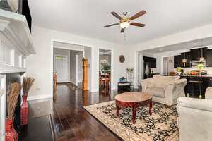 Spacious living room with dark wood floors, beige furniture, a patterned rug, a round wooden coffee table, ceiling fan, and open view into the kitchen and dining area. White trim and neutral walls throughout.
