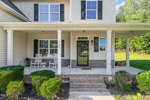 A house with beige siding, a brick front porch, black shutters, two white rocking chairs, and neatly trimmed bushes lining the walkway. The house number 1726 is displayed on a column next to the front door.