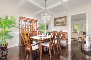 A dining room with a wooden table and eight chairs, a glass-front china cabinet, a chandelier, framed artwork on the wall, large potted plants, and dark wood floors. An adjoining living room is visible through an open doorway.