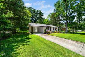 A single-story house with light-colored siding, a carport, and a concrete driveway, surrounded by green grass, trees, and landscaping under a partly cloudy sky.