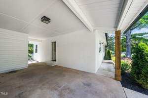 Covered carport area attached to a white brick house with a single door entry, concrete flooring, wooden support post, and some green shrubs visible along the perimeter.