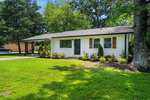 A small, single-story white brick house at 313 13th Street, Butner, with a dark front door, wooden shutters, a covered carport, and neatly trimmed shrubs in front, surrounded by green grass and tall trees.