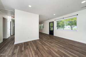 A spacious, empty room with white walls, wood-style flooring, large windows with blinds, a black door, and recessed ceiling lights. Natural light enters through the windows, and an open hallway leads to other rooms.