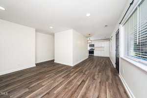 A spacious open-concept living area with wood-look flooring, white walls, recessed lighting, and large windows. The kitchen in the background features white cabinets, a stove, and a center light fixture.