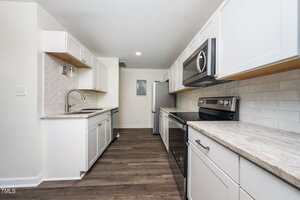A modern galley kitchen with white cabinets, stainless steel appliances, light stone countertops, a subway tile backsplash, and wood-look flooring. The space is clean and well-lit with neutral tones.