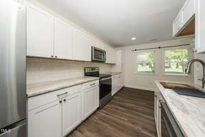 Modern kitchen with white cabinets, stainless steel appliances, light countertops, subway tile backsplash, dark wood floors, and two large windows letting in natural light.