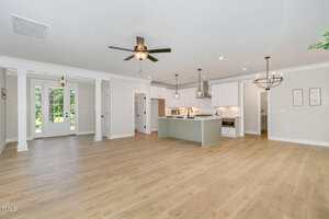 Open-concept living area with light wood flooring, white walls, ceiling fans, a kitchen with white cabinets and an island, two chandeliers, and double glass doors leading outside.
