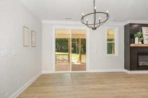 A bright, empty room with light wood flooring, white walls, a black chandelier, and double glass doors leading to a covered patio with a view of a green backyard. A modern fireplace is visible on the right.