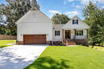 A white, two-story house at 503 Forest Road, Oxford, features brown wooden garage doors, a covered front porch, brick accents, and a sloped driveway. Neatly trimmed grass and trees surround the home beneath a blue sky with scattered clouds.