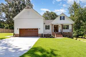 A white, two-story house at 503 Forest Road, Oxford, features brown wooden garage doors, a covered front porch, brick accents, and a sloped driveway. Neatly trimmed grass and trees surround the home beneath a blue sky with scattered clouds.