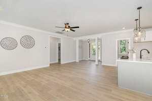 A spacious, unfurnished open-concept living area with light wood floors, white walls, a ceiling fan, two round wall decorations, and a modern kitchen with pendant lights and a view of greenery through large windows.