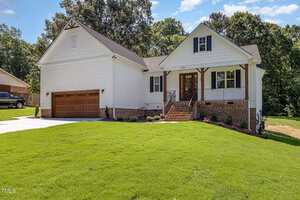 A white, single-family house with a gabled roof, brick accents, a wooden double garage door, front steps with a black railing, and a neatly mowed lawn, surrounded by trees under a blue sky.