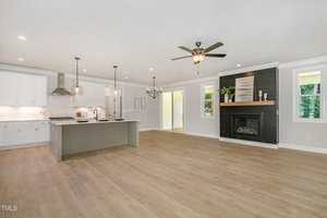 A spacious open-concept kitchen and living area with light wood flooring, white cabinets, a large island, stainless steel appliances, and a black fireplace with wood mantle. Ceiling fan and large windows provide natural light.