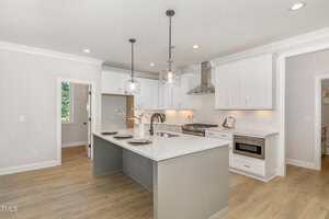 Modern kitchen with white cabinets, stainless steel appliances, a large gray island with bar stools, pendant lights, and light wood flooring. A window and a door are visible in the background.