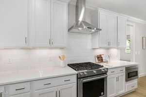 Modern kitchen with white cabinets, stainless steel gas stove and oven, range hood, white tile backsplash, and a built-in microwave. Countertops are clear except for a utensil holder and a cookbook stand.