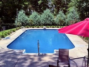 Rectangular outdoor swimming pool with metal ladders at both ends, surrounded by concrete decking. There is a red patio umbrella and two chairs in the foreground, with trees and shrubs in the background.