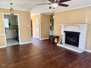 A living room with wood flooring, beige walls, a ceiling fan, a white fireplace with a black insert, and a small wooden bench. The room connects to a kitchen and a hallway, with natural light coming in.