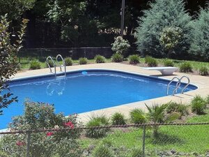 Rectangular outdoor swimming pool with metal ladders at both ends, a diving board, and surrounding landscaping, including bushes, small trees, and a chain-link fence in the foreground.