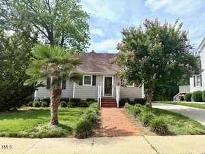 A single-story house with light-colored siding, a gable roof, and a red brick walkway leading to the front door. The yard has a palm tree, a flowering tree, shrubs, and a driveway on the right.