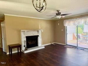 A living room with brown walls and dark wood floors features a white marble fireplace, a ceiling fan, a small table, and sliding glass doors with sheer curtains leading to a deck outside.