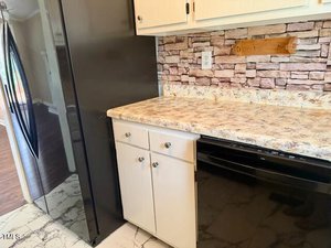 A kitchen corner with a black refrigerator, light-colored cabinets and countertop, faux stone backsplash, and a black dishwasher. The floor has a white marble pattern. A wooden utensil holder is mounted on the backsplash.