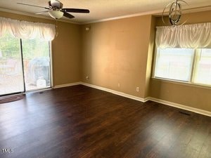 A mostly empty room with tan walls, dark wood flooring, two windows with white curtains, a ceiling fan, and sliding glass doors leading outside. A grill is visible on the patio through the glass doors.