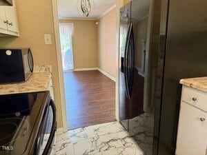 A view from a kitchen with black appliances and marble-style tile flooring, looking into a dining area with wood flooring, beige walls, and a window with sheer curtains.