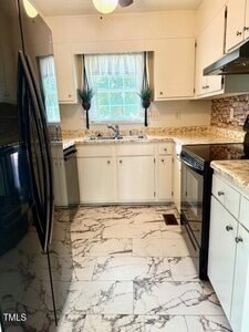 A small kitchen with white cabinets, marble-patterned floor, black appliances, and a window above the sink with white curtains. Countertops are beige with a stone-like backsplash. Two plants decorate the windowsill.