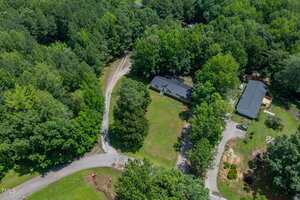 Aerial view of two homes surrounded by dense trees, each with a driveway leading to the street. The houses are separated by a large grassy area and patches of woodland. Several vehicles are parked outside one of the homes.