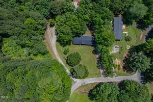 Aerial view of a house surrounded by trees with a curved driveway leading to the property. The yard is mostly grass with some landscaped areas and adjoining wooded sections. Another building is visible nearby.