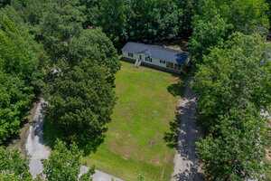 Aerial view of a single-story house with a dark roof, centered on a large grassy yard, surrounded by trees and a curved gravel driveway, on a sunny day.
