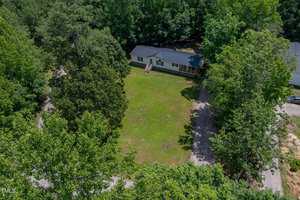 Aerial view of a single-story house with a dark roof and front steps, situated on a large grassy lawn surrounded by dense green trees and a gravel driveway.