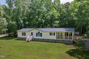 A single-story manufactured home at 40 Raccoon Crossing in Henderson features a dark metal roof, white siding, brick foundation, front steps, and a covered porch on a grassy lot surrounded by tall trees.