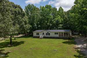 A single-story manufactured home with light-colored siding and a dark roof sits on a large grassy lawn, surrounded by tall, leafy trees under a partly cloudy sky. A wooden porch leads to the front entrance.