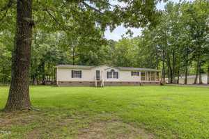 Single-story manufactured home with beige siding and a gable roof, set on a brick foundation. The house has a small front porch, steps leading to the front door, and is surrounded by green grass and trees.