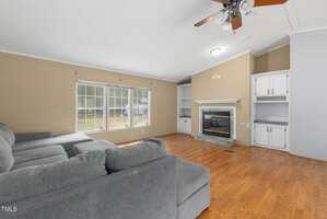 A living room with wood flooring, a gray sectional sofa, large windows, a fireplace with a mantel, built-in shelves on either side, and a ceiling fan with lights.
