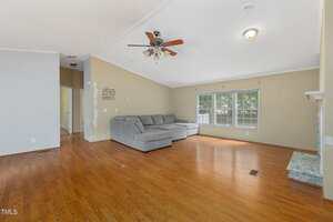 A spacious living room with light brown wood flooring, a gray sectional sofa, ceiling fan with lights, large windows letting in natural light, and beige walls with some visible paint patches.