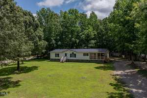 A single-story manufactured home with a covered front porch sits on a large grassy lot surrounded by trees. A gravel driveway leads up to the house, and sunlight filters through partly cloudy skies.
