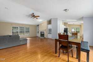 Open-concept living and dining area with wood flooring, a gray sectional sofa, fireplace, ceiling fan, dining table with chairs, built-in cabinetry, and large windows letting in natural light.