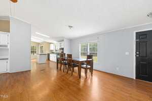 A dining area with a wooden table and four chairs sits on hardwood flooring. Large windows provide natural light. The space opens into a kitchen with white cabinets and visible appliances. The front door is black.