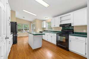 Kitchen with hardwood floors, white cabinets, green countertops, black appliances including a stove and refrigerator, an island in the center, and large windows letting in natural light.