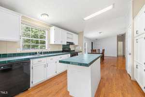 A kitchen with white cabinets, a green tiled countertop and island, black appliances, and wood flooring. A large window over the sink lets in natural light. The dining area is visible in the background.
