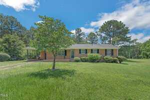 Single-story brick house with white trim, covered carport, large front lawn, small tree in the yard, and several bushes. Surrounded by tall pine trees under a mostly blue sky with some clouds.