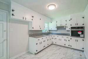 A kitchen with white cabinets, black hardware, granite countertops, a stainless steel sink, a built-in oven, vent hood, and marble-patterned floor tiles. A window is above the sink, and a round ceiling light is on.