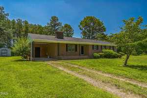 Single-story brick house with a covered carport, chimney, and black shutters. The house sits on a grassy lawn with trees in the background and a gravel driveway leading to the carport.