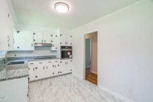 A kitchen with white cabinets, silver handles, granite countertops, built-in oven, stainless steel range hood, and marble-patterned tile floor. A doorway leads to a room with wood flooring.