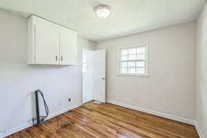 A small empty room with light-colored walls, a wooden floor, a window, a door leading to another room, a white ceiling light, a wall-mounted cabinet, and exposed plumbing and electrical outlets on one wall.
