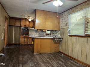 A kitchen with wood cabinets, a built-in oven, wood-paneled walls, floral wallpaper, and brown tile flooring. A small breakfast bar divides the kitchen from the dining area. A window has sheer white curtains.
