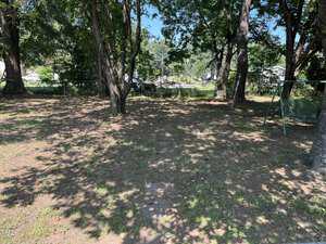 A backyard with patchy grass and dirt, scattered shade from several large trees, a swing set frame on the right, and some cars and houses visible in the background.